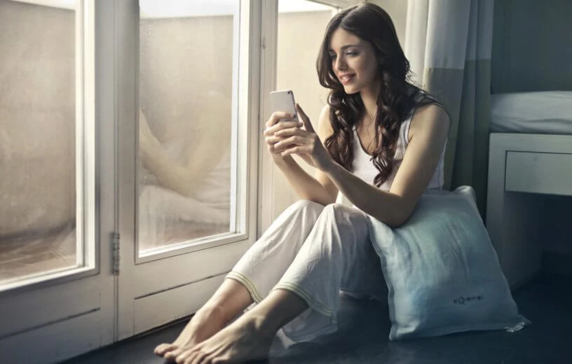A woman using her phone while sitting beside a bright window