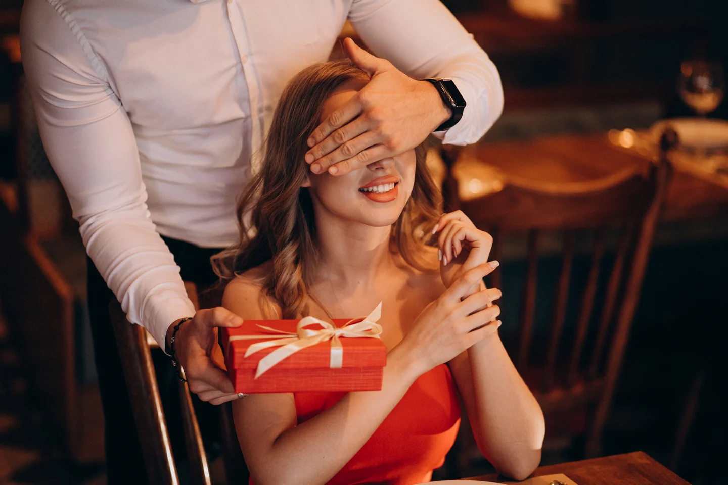 A woman smiling as a man surprises her with a wrapped gift